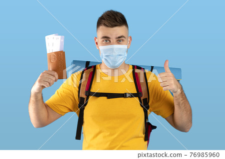Young backpacker in face mask holding international passport with plane tickets and showing thumb up on blue background Young backpacker in face mask holding international passport with plane tickets and showing thumb up on blue background 76985960