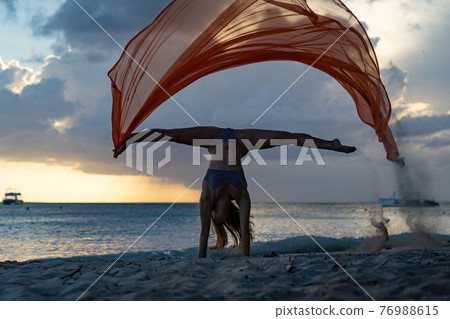 Silhouette of flexible fit woman doing handstand with silk during dramatic sunset with stormy clouds on the seascape background. Concept of happiness, freedom and carefree. Silhouette of flexible fit woman doing handstand with silk during dramatic sunset with stormy clouds on the seascape background. Concept of happiness, freedom and carefree. 76988615