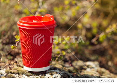 red cardboard glass for coffee is standing on a stone, outdoors, against the background of fresh greenery, walking or picnic in nature, concept 76990045