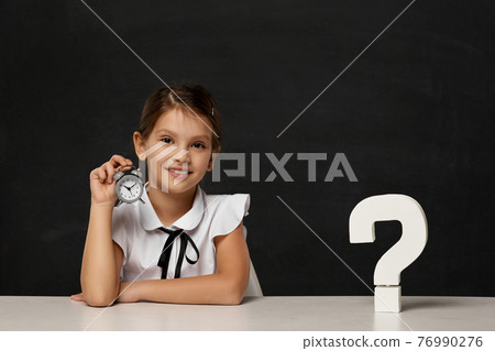 happy schoolgirl with alarm clock sitting at table 76990276