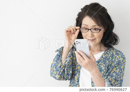 A smiling woman in her 60s wearing reading glasses and looking at her smartphone 76990627
