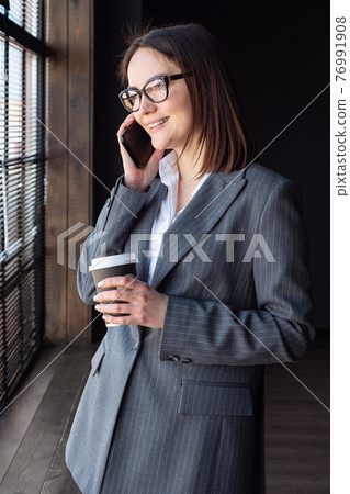 Business woman with coffee stands by the window and speaks on the phone Business woman with coffee stands by the window and speaks on the phone 76991908