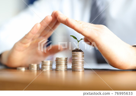 Businessman holds his hands with house above cylinders of coins Businessman holds his hands with house above cylinders of coins 76995633