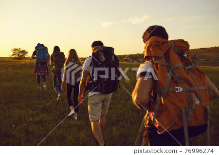 Group of travelers with rucksacks walking in single file in the field during active summer vacation 76996274