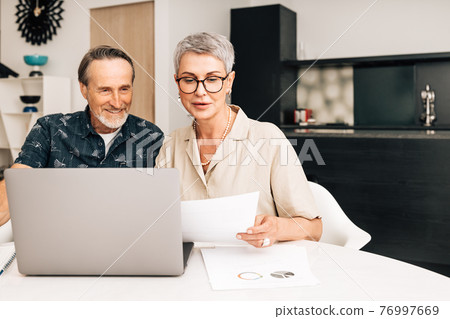 Senior retired couple at a table in dining room using a laptop computer. Two mature people looking at paperwork and discussing their finances. Senior retired couple at a table in dining room using a laptop computer. Two mature people looking at paperwork and discussing their finances. 76997669