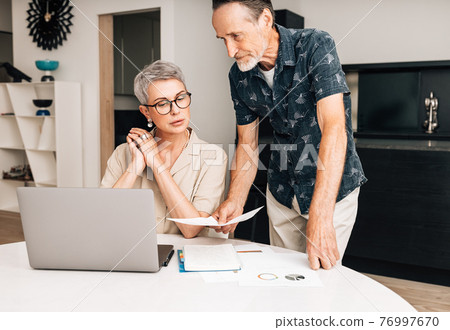 Senior couple with laptop calculating finances in the dining room. Two retired people with documents at home. Senior couple with laptop calculating finances in the dining room. Two retired people with documents at home. 76997670