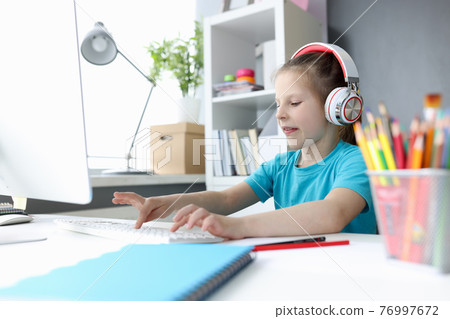 Little girl in headphones in front of computer is typing on keyboard Little girl in headphones in front of computer is typing on keyboard 76997672