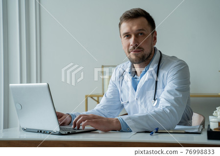 Doctor using laptop in medical office. Man is working with documents and laptop in his medical office 76998833