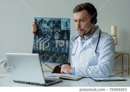 Male doctor listening to patient during telemedicine session. Man doctor having online consultation on laptop holding x-ray image and discussing diagnosis Male doctor listening to patient during telemedicine session. Man doctor having online consultation on laptop holding x-ray image and discussing diagnosis 76998863