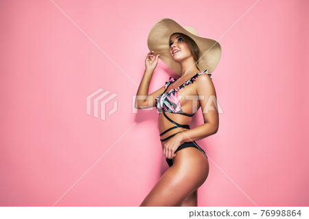 A gorgeous trendy fit woman in swimwear and hat while she enjoying rest with sunlight isolated over pink background. Travel, tourism, journey, trip, tour, summer concept 76998864
