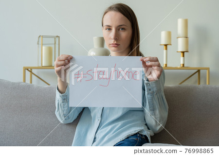 A woman holding a piece of paper with the inscription STAY HOME is sitting on the sofa A woman holding a piece of paper with the inscription STAY HOME is sitting on the sofa 76998865