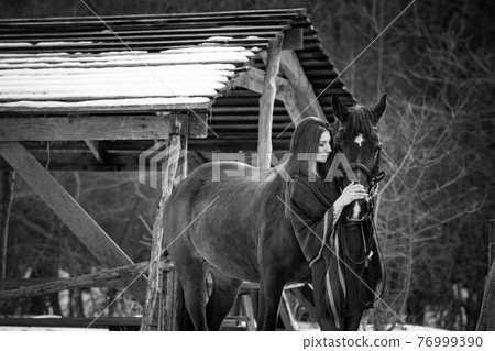 Beautiful girl hugs a horse near wooden buildings on a winter day, black and white photography Beautiful girl hugs a horse near wooden buildings on a winter day, black and white photography 76999390