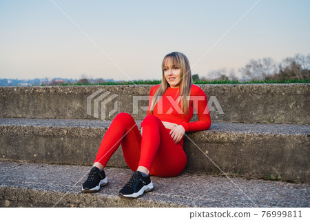 Portrait of a smiling confident fitness woman in red sport wear resting after work out or running outdoors on sunset. Jogging in the stadium. Active lifestyle and health care. Copy space. Portrait of a smiling confident fitness woman in red sport wear resting after work out or running outdoors on sunset. Jogging in the stadium. Active lifestyle and health care. Copy space. 76999811