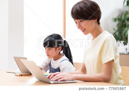 Girl learning online next to her mother teleworking at the kitchen table 77005867