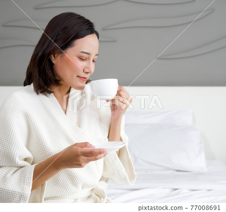 Asian woman in a bathrobe enjoys having morning tea on bed. Asian woman in a bathrobe enjoys having morning tea on bed. 77008691