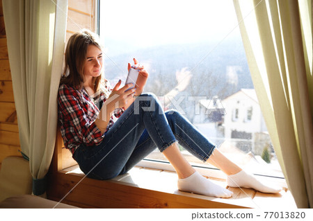 young girl talking on the phone while sitting by the window 77013820