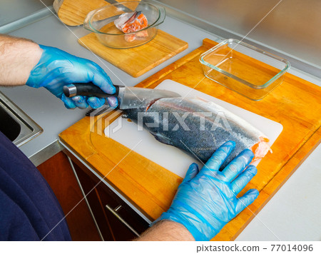 The hands of the cook in gloves cut the body of the salmon with a knife. 77014096