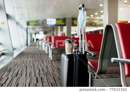 Next to the seats in an empty airport is a suitcase, on it is a glass of coffee, hanging a medical protective mask. Coronavirus, social distance, isolation 77015302