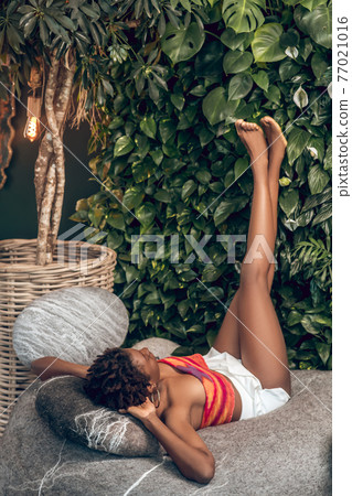 Dark-skinned young woman in a bright top and white shorts laying with her legs up Dark-skinned young woman in a bright top and white shorts laying with her legs up 77021016