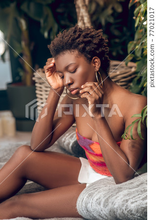 Dark-skinned young woman in a bright top and white shorts looking thoughtful Dark-skinned young woman in a bright top and white shorts looking thoughtful 77021017