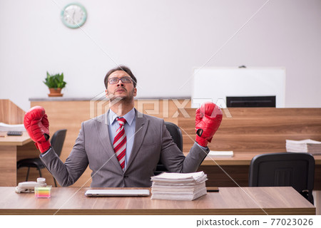 Young male employee wearing boxing gloves at workplace Young male employee wearing boxing gloves at workplace 77023026