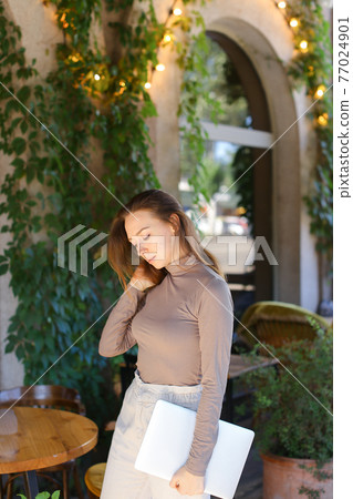 Young caucasian girl holding laptop, standing near green plants and window in street at cafe. Young caucasian girl holding laptop, standing near green plants and window in street at cafe. 77024901