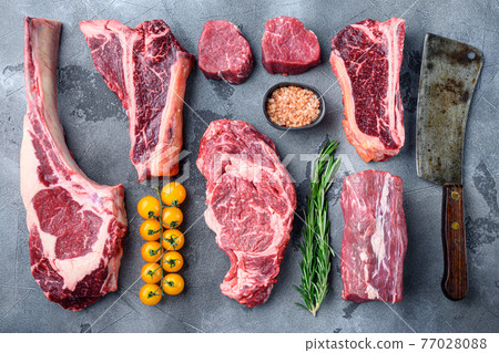 Variety of raw beef meat steaks for grilling with seasoning and utensils, tomahawk, t bone, club steak, rib eye and tenderloin cuts, on gray stone background, top view flat lay Variety of raw beef meat steaks for grilling with seasoning and utensils, tomahawk, t bone, club steak, rib eye and tenderloin cuts, on gray stone background, top view flat lay 77028088