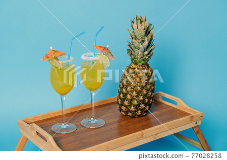 Pineapple and two glasses with cocktails decorated with cocktail umbrella on bamboo serving tray Pineapple and two glasses with cocktails decorated with cocktail umbrella on bamboo serving tray 77028258