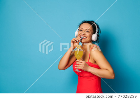 Charming woman with beautiful smile wearing headphones posing to camera with a cocktail in hands Charming woman with beautiful smile wearing headphones posing to camera with a cocktail in hands 77028259