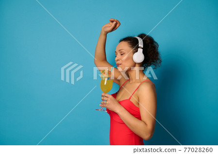 Brunette woman in a red swimsuit drinking juice from a straw and enjoying listening to music with headphones. 77028260