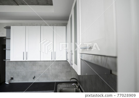 Modern minimalism style kitchen interior in monochrome tones. Custom kitchen with grey white facadesmdf and dark grey countertop with installed kitchen hood and sink. Modular kitchen from chipboard. 77028909