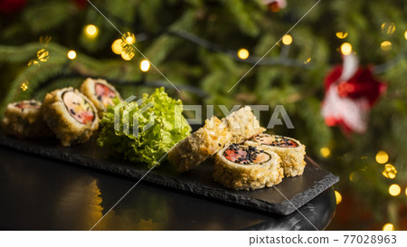 Custom sushi roll in tempura with nori, fresh salmon, tuna, avocado, masago caviar, drizzled with pineapple sauce with salad pouring as decoration on a black plate on a wooden table for Christmas. 77028963