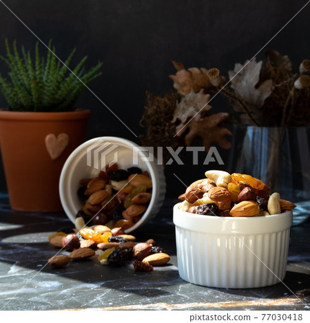 Mix of delicious assorted nuts and dried fruit in white cups on a table, sunny morning. Healthy gourmet vegetarian natural snack. 77030418