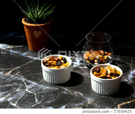 Mix of delicious assorted nuts and dried fruit in white cups on a table, sunny morning. Healthy gourmet vegetarian natural snack. 77030425