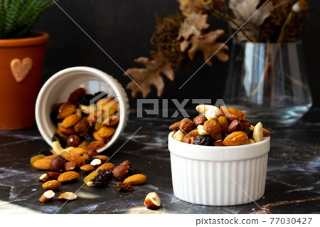Mix of delicious assorted nuts and dried fruit in white cups on a table, sunny morning. Healthy gourmet vegetarian natural snack. 77030427