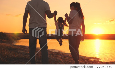 A small child holds the hand of mom and dad at the sunset sky. Childhood with parents at dawn. Happy family life. Mother, father and kid in the glare of the sun. Travel along the beach, near the coast 77030530
