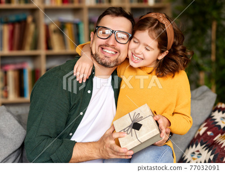 Excited girl daughter giving gift box for young loving father on holiday while sitting together on sofa 77032091