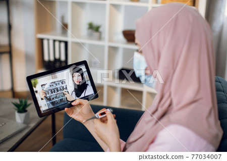 Side blurred view of sick young Muslim woman holding thermometer in hands, doing a medical consultation online with female Arabian gp doctor on digital tablet at home. Side blurred view of sick young Muslim woman holding thermometer in hands, doing a medical consultation online with female Arabian gp doctor on digital tablet at home. 77034007