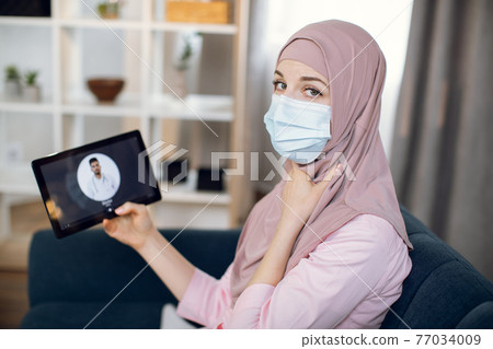 Coronavirus pandemic. Sick Muslim woman in hijab and mask, suffering from sore throat, using tablet pc and making video call with Indian male doctor while staying at home. Coronavirus pandemic. Sick Muslim woman in hijab and mask, suffering from sore throat, using tablet pc and making video call with Indian male doctor while staying at home. 77034009