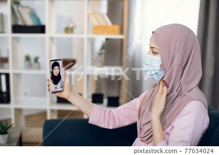 Muslim young woman patient with sore throat, having video chat with Arabian female doctor via smartphone app, sitting on sofa at home. Tele medicine, technology and healthcare concept Muslim young woman patient with sore throat, having video chat with Arabian female doctor via smartphone app, sitting on sofa at home. Tele medicine, technology and healthcare concept 77034024