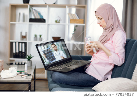 Remote doctor consultation online. Young sick Muslim woman in hijab takes an online consultation advice, talking with male doctor using laptop at home, showing him different blisters with pills 77034037