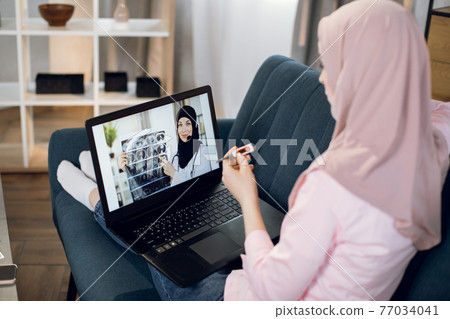 Pc monitor view over shoulder of young Muslim woman, sitting on sofa at home with thermometer . Young Arabian female doctor wear headset, gives consultation to patient about CT scan 77034041