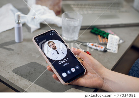 Remote consultation concept. Close-up of patient hand, holding smartphone, calling to male doctor for online medical device. Table with medicines, thermometer, glass and laptop. 77034081