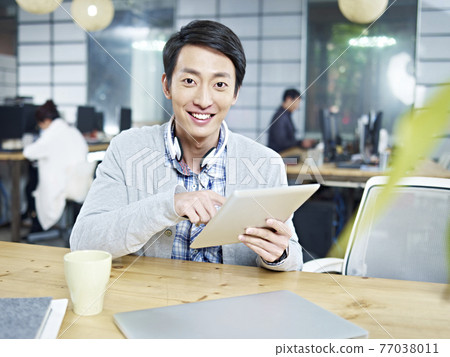 young asian business man sitting at desk in office holding tablet computer looking at camera smiling young asian business man sitting at desk in office holding tablet computer looking at camera smiling 77038011