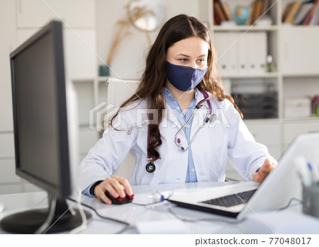 Woman doctor in protective mask sitting at workplace with computer in office Woman doctor in protective mask sitting at workplace with computer in office 77048017