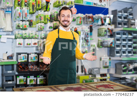 glad male seller showing assortment in garden equipment shop 77048237