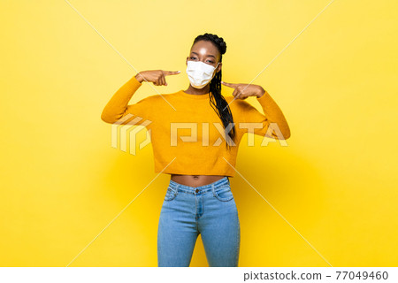 Young hygienic healthy African woman wearing and pointing to face mask Young hygienic healthy African woman wearing and pointing to face mask 77049460