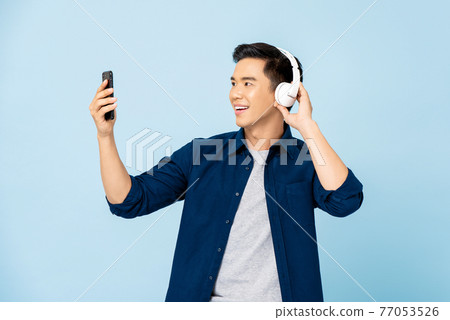 Happy smiling Asian man wearing headphones listening to music from mobile phone Happy smiling Asian man wearing headphones listening to music from mobile phone 77053526