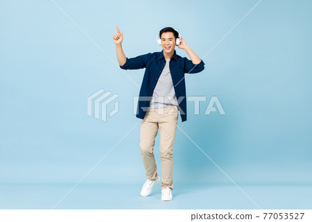 Happy smiling young handsome Asian man listening to music with wireless headphones pointing hand up 77053527