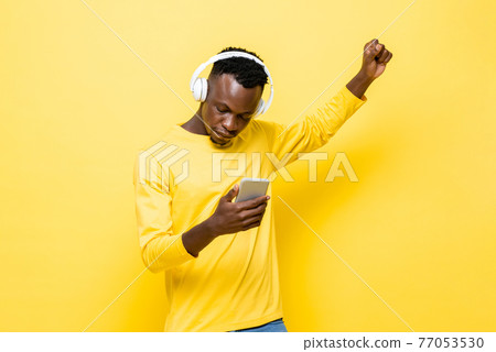 Young African man in casual attire wearing wireless headphones listening to music with mobile phone Young African man in casual attire wearing wireless headphones listening to music with mobile phone 77053530